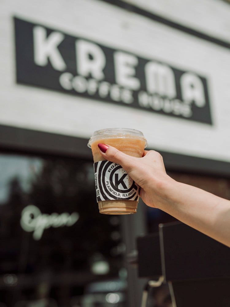 Iced latte held up in front of the Krema Coffee House sign